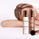 Close up of two aerosol cans of foundation and primer with a brush and compact. Behind the makeup products is a women laying her head on a surface. Image is light with soft pink tones.