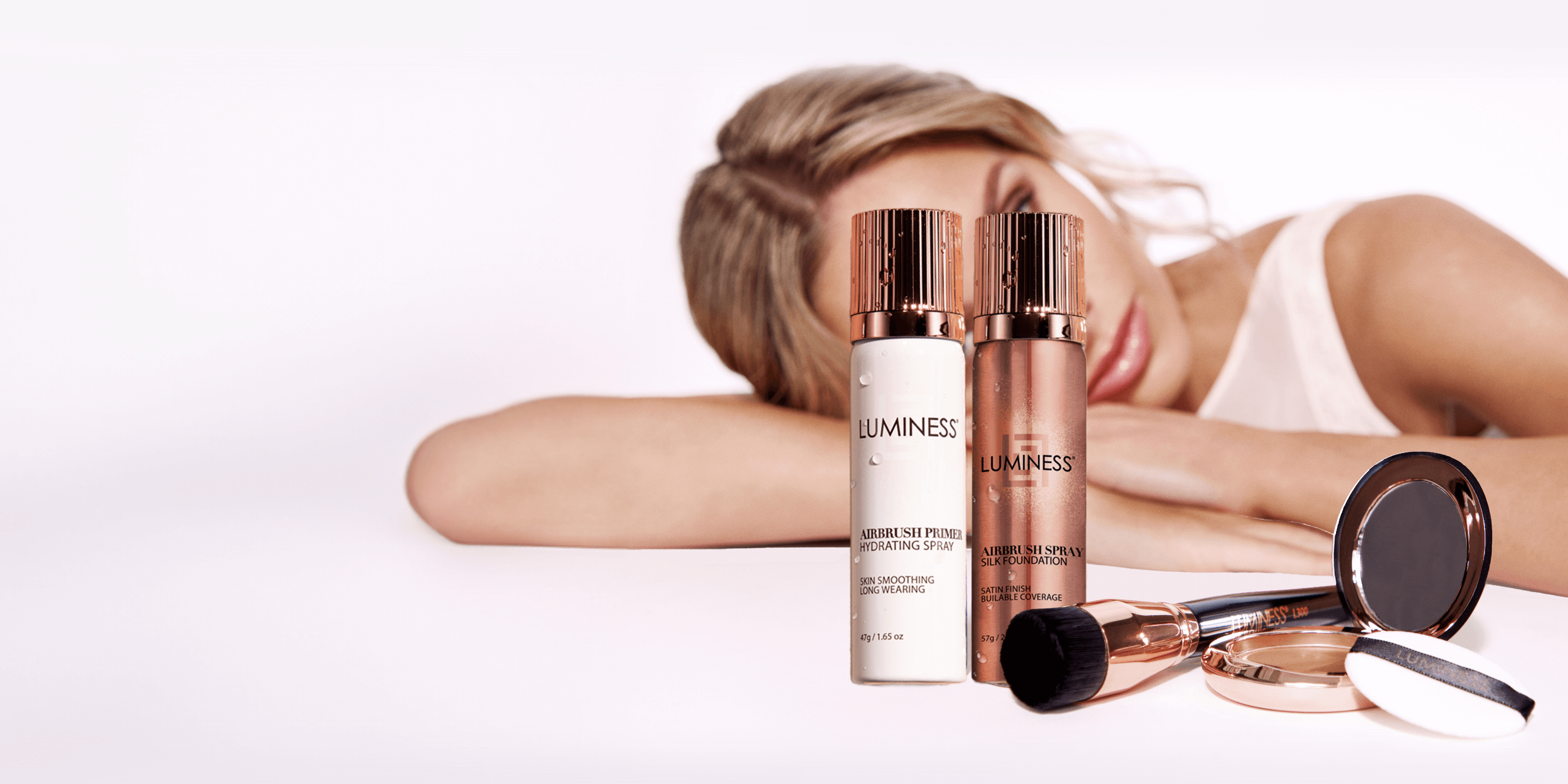 Close up of two aerosol cans of foundation and primer with a brush and compact. Behind the makeup products is a women laying her head on a surface. Image is light with soft pink tones.