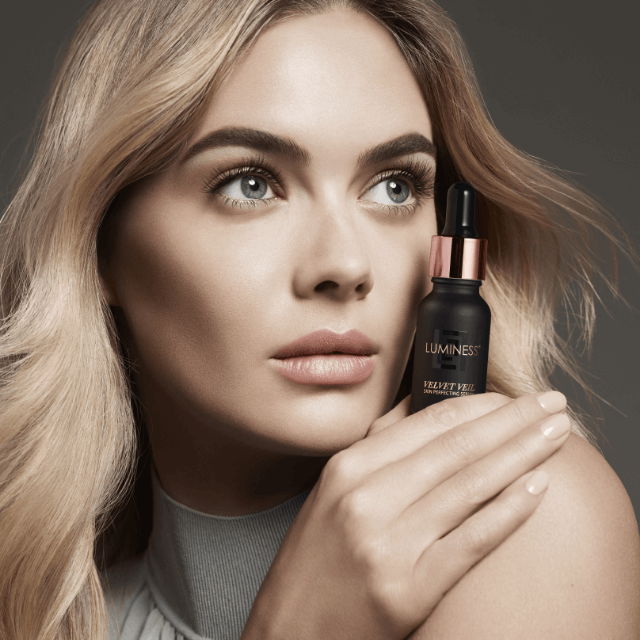 Close up photo of a woman's face as she holds a bottle of foundation serum