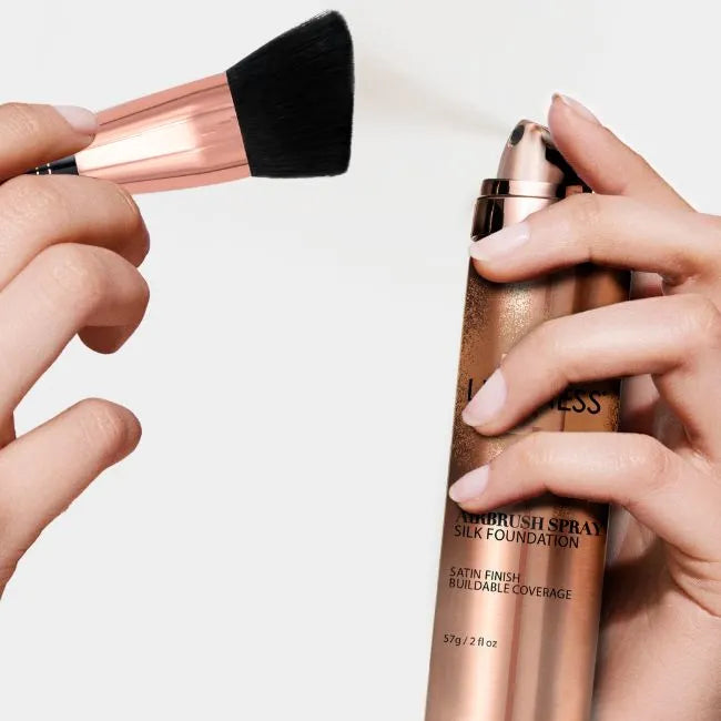 Photo of hands holding a makeup brush next to a bottle of spray foundation. Brush is being sprayed with foundation